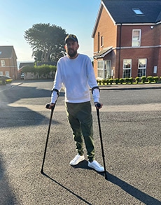 A light-skinned man wearing a hat, sweatshirt, pants and sneakers stands with crutches outsid. 
